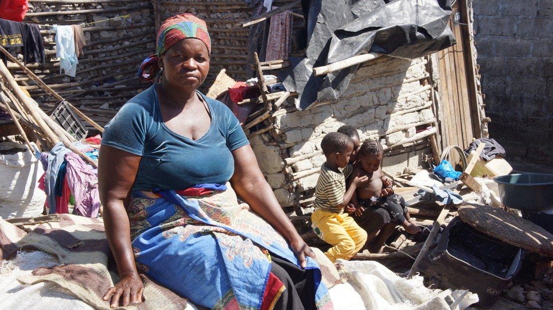 A woman sits amid rubble, gravel, bricks and plastic, staring into the camera with a closed face. Behind her, three small children huddle together.